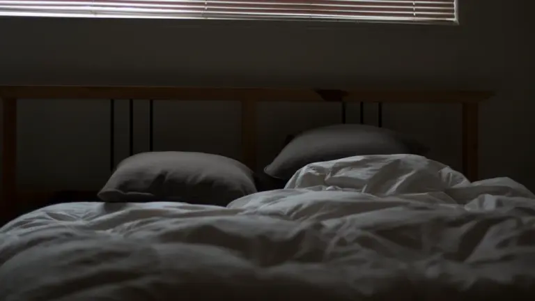 Ein ungemachtes Bett mit heruntergelassenen Jalousien bei abgedunkeltem, warmem Tageslicht im Schlafzimmer.