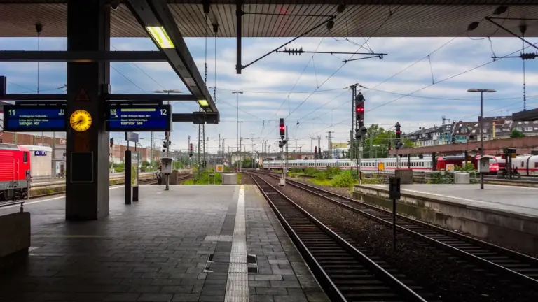 Ein typischer deutscher Bahnhofsbahnsteig mit Gleisen und digitalen Anzeigetafeln an einem bewölkten Tag.