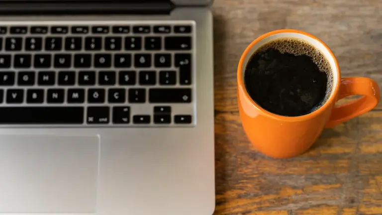 Ein Laptop steht neben einer orangefarbenen Tasse mit schwarzem Kaffee auf einem Holztisch.