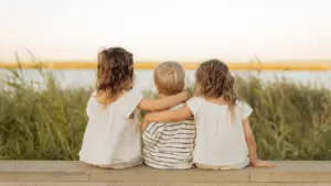 Drei Kinder sitzen nebeneinander an einem Gewässer und blicken gemeinsam in die Ferne.