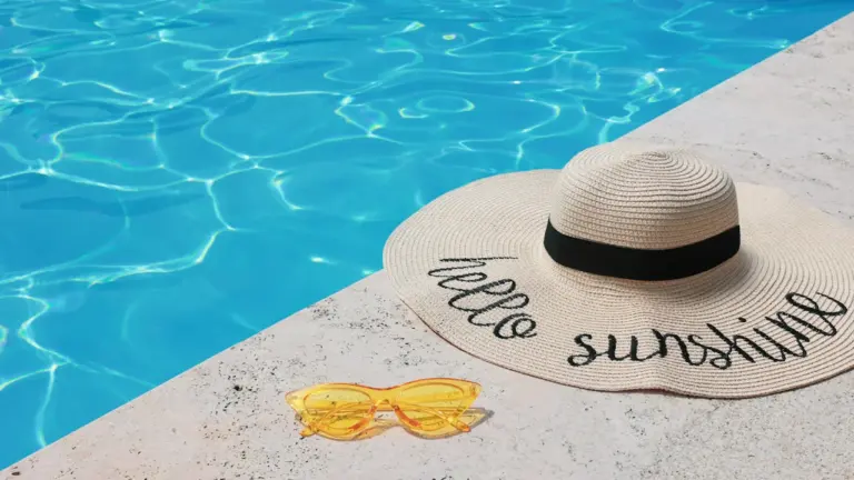Sonnenhut und gelbe Sonnenbrille am Rand eines blauen Swimmingpools bei strahlendem Sonnenschein.